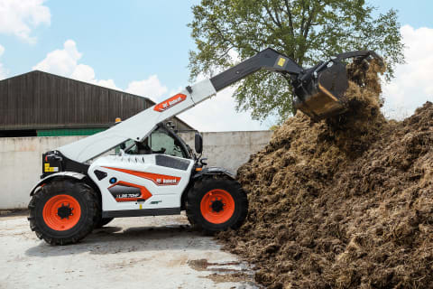 Ein Bobcat-Teleskop mit Schaufelgreifer und ausgefahrenem Löffelstiel kippt auf einem offenen Bauernhof Dung auf einen Haufen. 