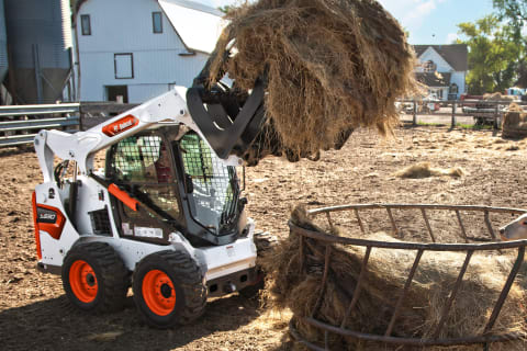 Оснащенный ковшом с захватом мини-погрузчик Bobcat с бортовым поворотом под управлением мужчины-оператора перевозит сено на ферме.