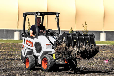 Un operatore al lavoro con una pala articolata di piccole dimensioni Bobcat con benna con pinza mentre trasporta terra su un terreno in piano accanto a un sito industriale.