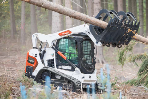 Ein männlicher Bediener, der einen Bobcat-Kompakt-Raupenlader mit ausgefahrenen Armen und dem Industriegreifer als Anbaugerät verwendet, um einen langen Baumstamm in einem Wald zu transportieren.