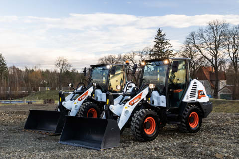 Twee compacte wielladers van Bobcat staan naast elkaar geparkeerd op een vlak grindterrein terwijl achter hen de zon ondergaat.