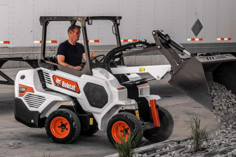 A male operator using a Bobcat Small Articulated Loader with the Utility Bucket attachment to dump small stones into a border near an industrial loading bay.