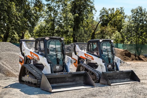 Dos cargadoras compactas de orugas Bobcat aparcadas una junto a la otra en una superficie plana de grava en un día soleado.