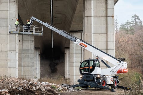 A Bobcat Rotary Telehandler with an outstretched boom arm raising two construction workers toward the underside of a large bridge. 