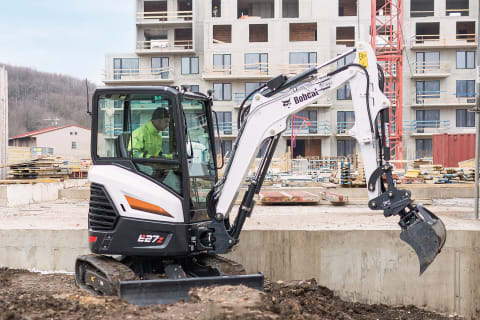 Ein männlicher Fahrer verwendet einen Bobcat-Minibagger, um Erde in der Nähe einer Betonmauer auf einer Wohnanlage zu bewegen.