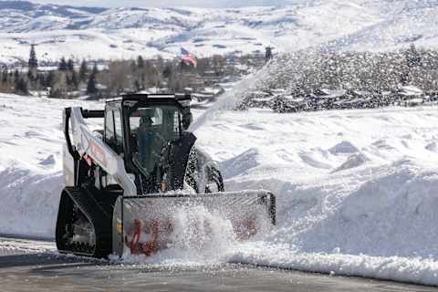 A Bobcat Compact Track Loader Clears Snow Along the Edge of a Parking Lot With a Snowblower Attachment