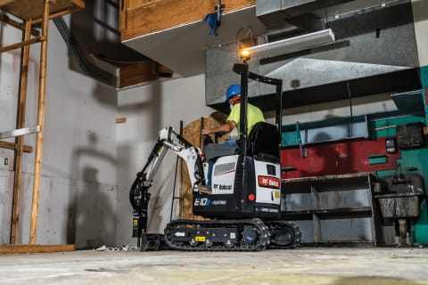 A Bobcat E10e battery-electric compact excavator uses a nitrogen breaker on a concrete floor inside an enclosed room.