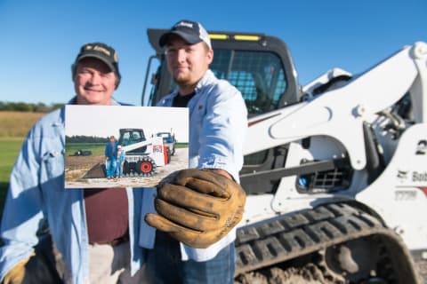 Phil Sylvester and his son Tyler standing by their Bobcat T770 Compact Track Loader, with Tyler holding an old photograph of them with their first Bobcat Machine an 863 Skid-Steer Loader