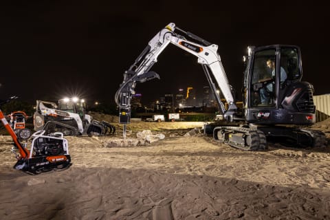 A Large Excavator, Surrounded by Various Bobcat Machines, Uses a Breaker Attachment on Jobsite at Night