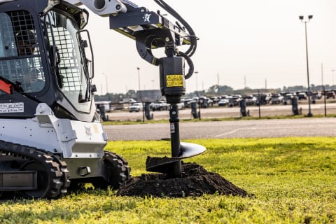 A Bobcat compact track loader uses its auger attachment to dig a hole in grass. 