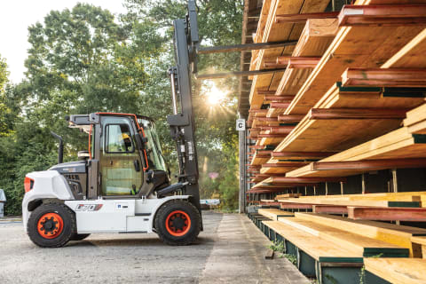 A Bobcat forklift lifts wood planks from a shelf at an outdoor lumber yard. 