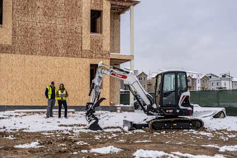 A Bobcat Compact Excavator Is Parked on the Snowy Site of a Building Construction. Two Workers in Safety Vests Talk Nearby.