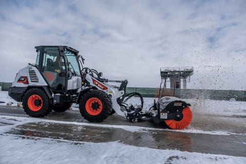 A Female Operator Uses a Bobcat Compact Wheel Loader Equipped With an Angle Broom Attachment to Remove Snow From a Roadway