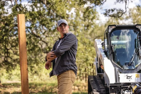 A Man Rolls Up His Sleeves While Standing Near a Recently Placed Fence Post. A Bobcat Compact Track Loader Is Parked in the Background.