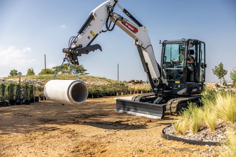 A Bobcat E60 Compact Excavator Lifts a Large Concrete Pipe Hanging From a Chain