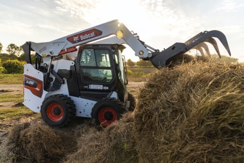 A Bobcat S86 Skid-Steer Loader Bites Into a Large Hay Bale With an Industrial Grapple Attachment