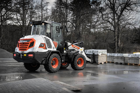 An outdoor action shot of Bobcat's L75 compact wheel loader with a male operator transporting a pallet of bricks on a grey, rainy day. 