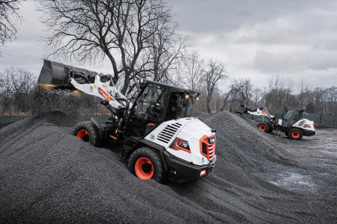 An outdoor action shot of Bobcat L75 and L85 compact wheel loaders with male operators transporting buckets of gravel on a grey, rainy day.