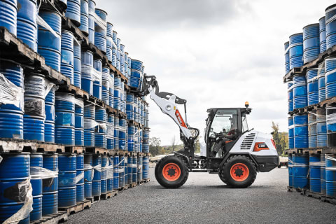 An outdoor action shot showcasing the side of Bobcat's L85 compact wheel loader and a male operator move barrels.