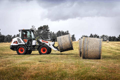 An outdoor action shot of a L95 Compact Wheel Loader with a male operator, transporting a bale of hay on a meadow on a cloudy, autumn day.