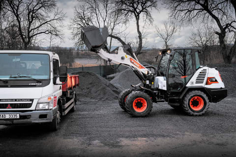 Actiefoto buiten van de L75 compacte wiellader die op een bewolkte dag een vrachtwagen vanaf de zijkant met grind laadt met behulp van een bak.