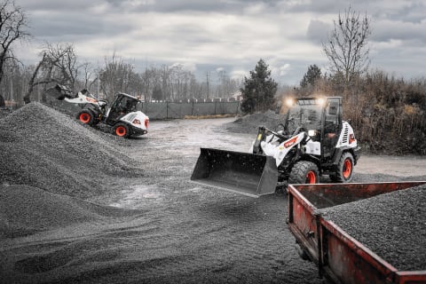 An outdoor action shot of L75 and L85 Compact Wheel Loaders loading and transporting gravel on a cloudy, sleety day.