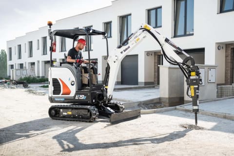 Photo extérieure en action d’un opérateur utilisant la machine E19e avec l’accessoire marteau hydraulique pour faire des travaux sur une route.