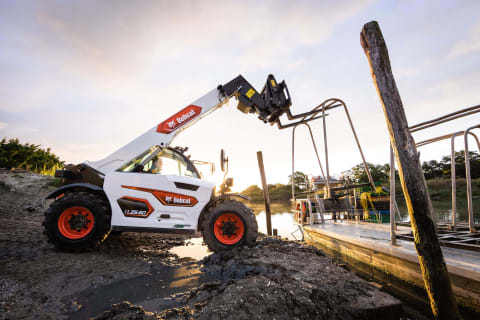 Eine Aufnahme im Freien eines männlichen Fahrers in Action, der mit dem kompakten Teleskopen TL25.60 von Bobcat einen Metallpfahl am Ufer eines Flusses anhebt.