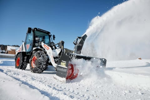 A male operator operates a Bobcat L95 compact wheel loader equipped with a snowblower attachment as it clears a snow-covered area. 