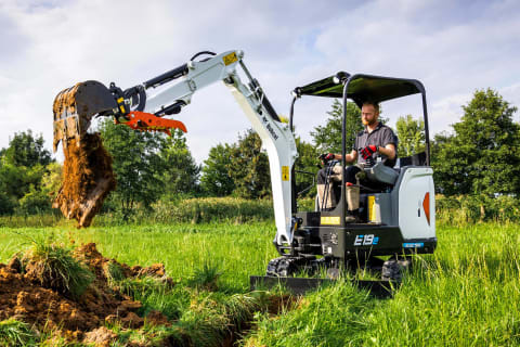 Image of a male operator expertly operating Bobcat's E19e excavator to dig up dirt in a lush grassy field. 