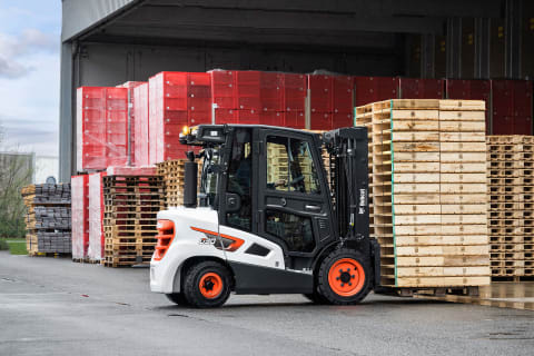 An outdoor action shot of a male operator using a Bobcat diesel forklift to transport wood pallets in a warehouse setting.