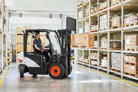 An indoor action shot of a male operator using a Bobcat diesel forklift in a warehouse, carefully moving a box labeled "handle with care".