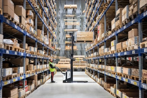 An indoor side view of a Bobcat BSL20S-7 with a skilled female operator navigating down the aisle of a well-lit and organized warehouse.