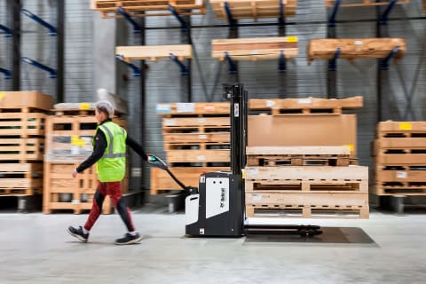 An indoor action shot from the side, featuring a skilled female operator maneuvering a Bobcat BSL16S-7 down a well-lit and organized warehouse aisle.