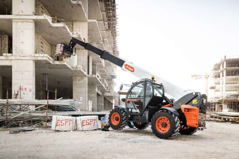 Динамичный натурный снимок телескопического погрузчика Bobcat T40.180SLP с разложенной стрелой под управлением мужчины-оператора, подающего материалы на второй этаж строящегося здания.