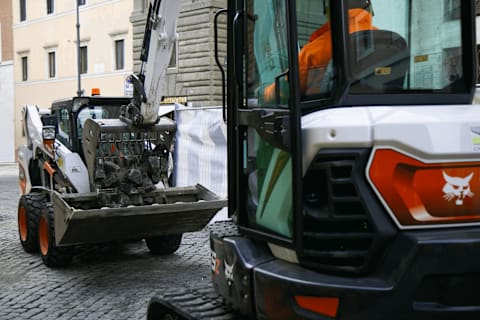 A close-up daytime shot of two Bobcat machines and their operators working on a cobblestone street.