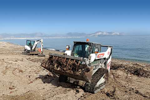 Two Bobcat machines on the rocky, sandy shore of Bagheria Beach, with the sea and mountains in the background.