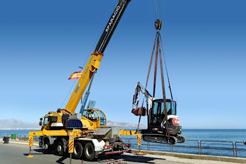 Una grúa Sparacio elevando una miniexcavadora Bobcat E35z con un impresionante paisaje de mar y montaña de fondo.