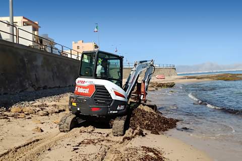 Ein männlicher Fahrer, der mit einem Bobcat-Minibagger E35z und einer Schaufel als Anbaugerät an einem Sandstrand Sand schaufelt, mit einem Dorf, dem Meer und den Bergen im Hintergrund.