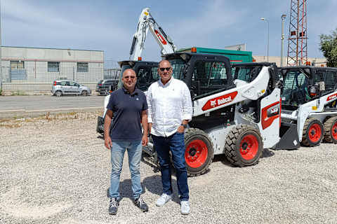 Twee lachende mannen met zonnebril staan op een zonnige dag voor een Bobcat S660-schranklader.