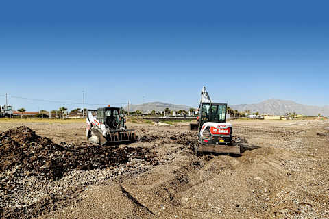 Two Bobcat machines with one male operator working on a rocky, sandy landscape with mountains in the background.