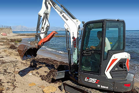 Operador utilizando una miniexcavadora Bobcat E35z con un implemento de cucharón en la playa de arena de Bagheria, con el mar y las montañas de fondo.