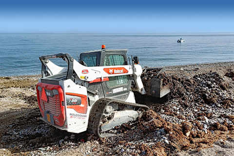 Eine Profilaufnahme eines Bobcat T66 am felsigen sandigen Ufer des Sandstrands von Bagheria, mit dem schönen Meer im Hintergrund.