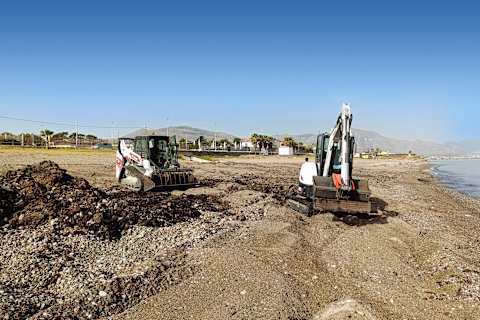 Two Bobcat machines working on a rocky, sandy landscape with mountains in the background.