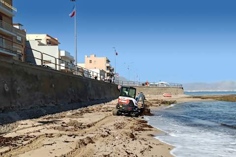 Une mini-pelle E35z Bobcat évoluant sur une plage de sable avec un village, la mer et des montagnes en arrière-plan.