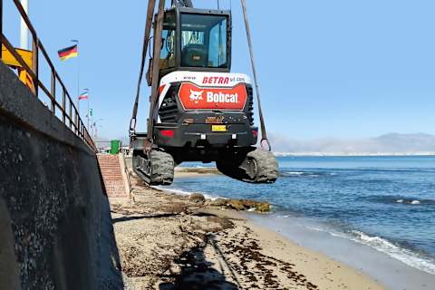 Een Sparacio-kraan laat een Bobcat E35z-minigraafmachine zakken op het zandstrand van Bagheria, met de zee en bergen op de achtergrond. 