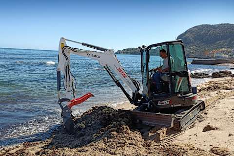 Мини-экскаватор Bobcat E35z под управлением мужчины-оператора просеивает песок в ковше на берегу пляжа, а вокруг набегают волны.