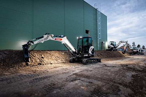 Two male operators working with Bobcat excavators, E55z and E50Z, at night in front of a sage green building.