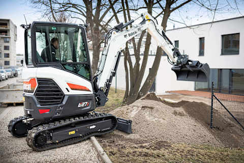 A profile shot of a male operator using a Bobcat E27z excavator with a bucket attachment to scoop dirt in front of an apartment building.