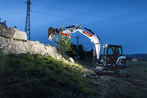 Scatto notturno di profilo di un operatore che utilizza un escavatore Bobcat E88 per togliere i detriti di un muro crollato.